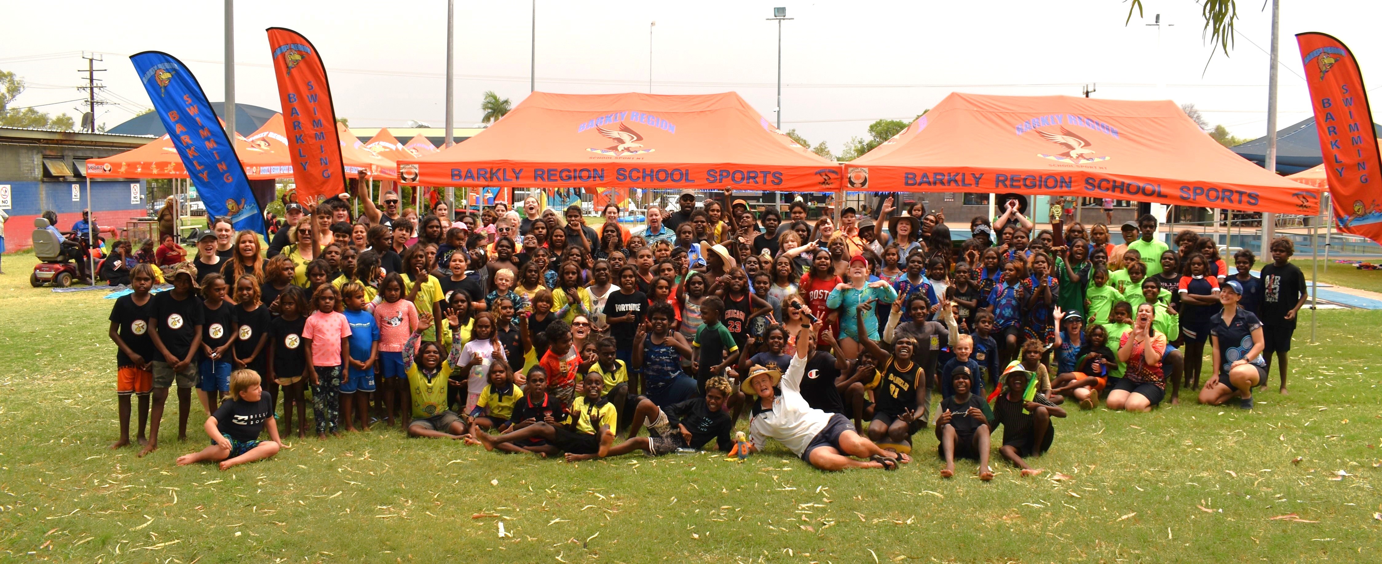 A large group of Indigenous students and a couple of teachers in front of orange marquees labelled 'Barkly Region School Sports'