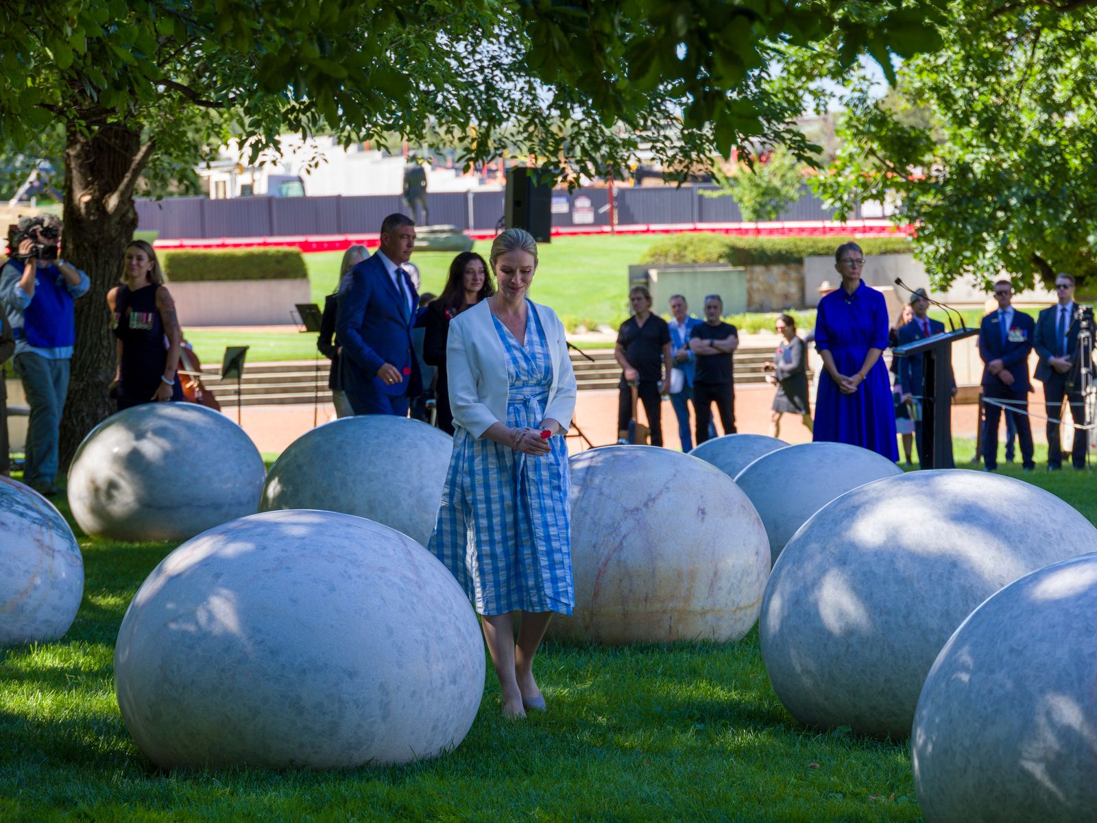 A woman in a white and blue checked dress stands on grass among marble boulders