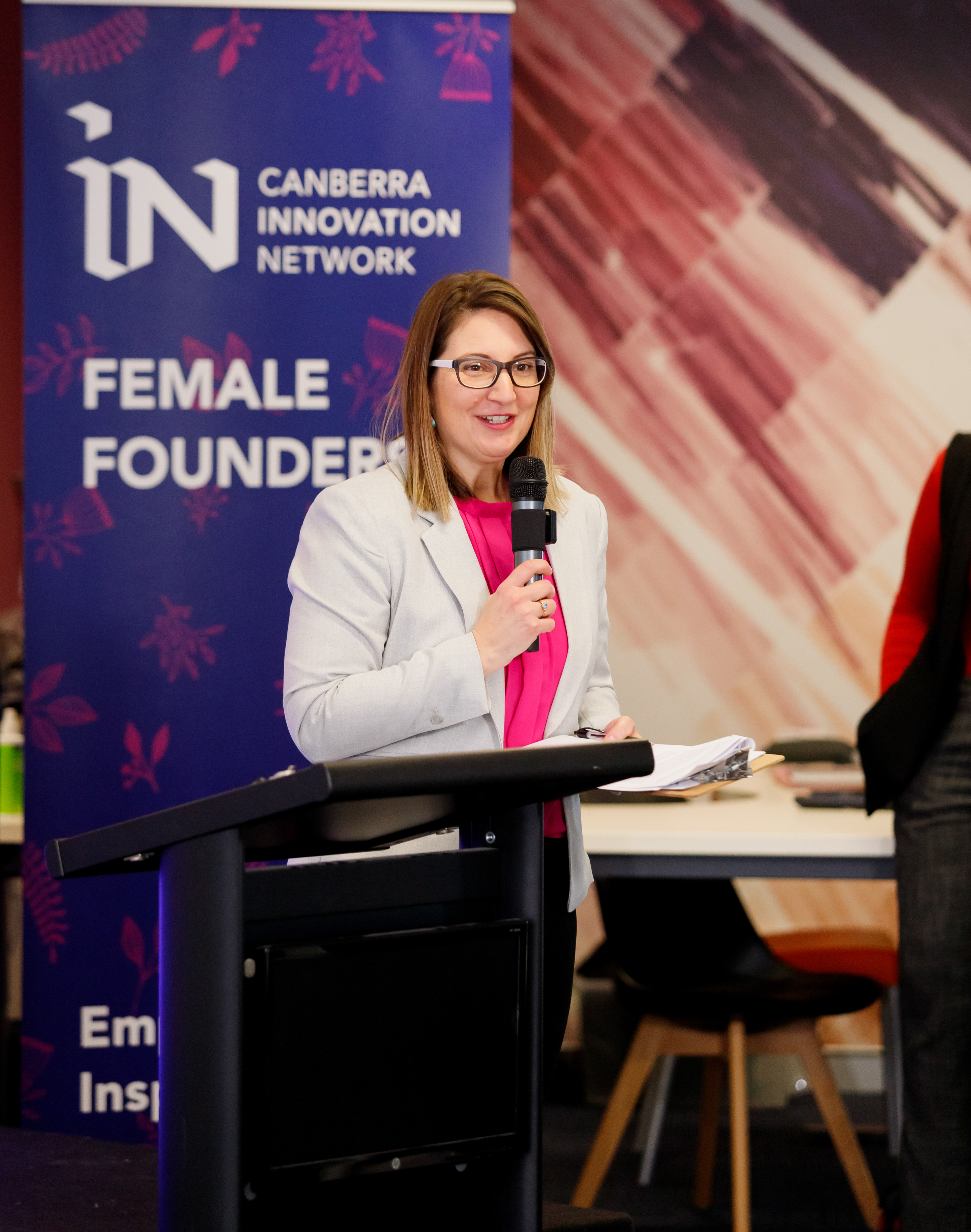 A woman in a white blazer and pink shirt speaks from a lectern