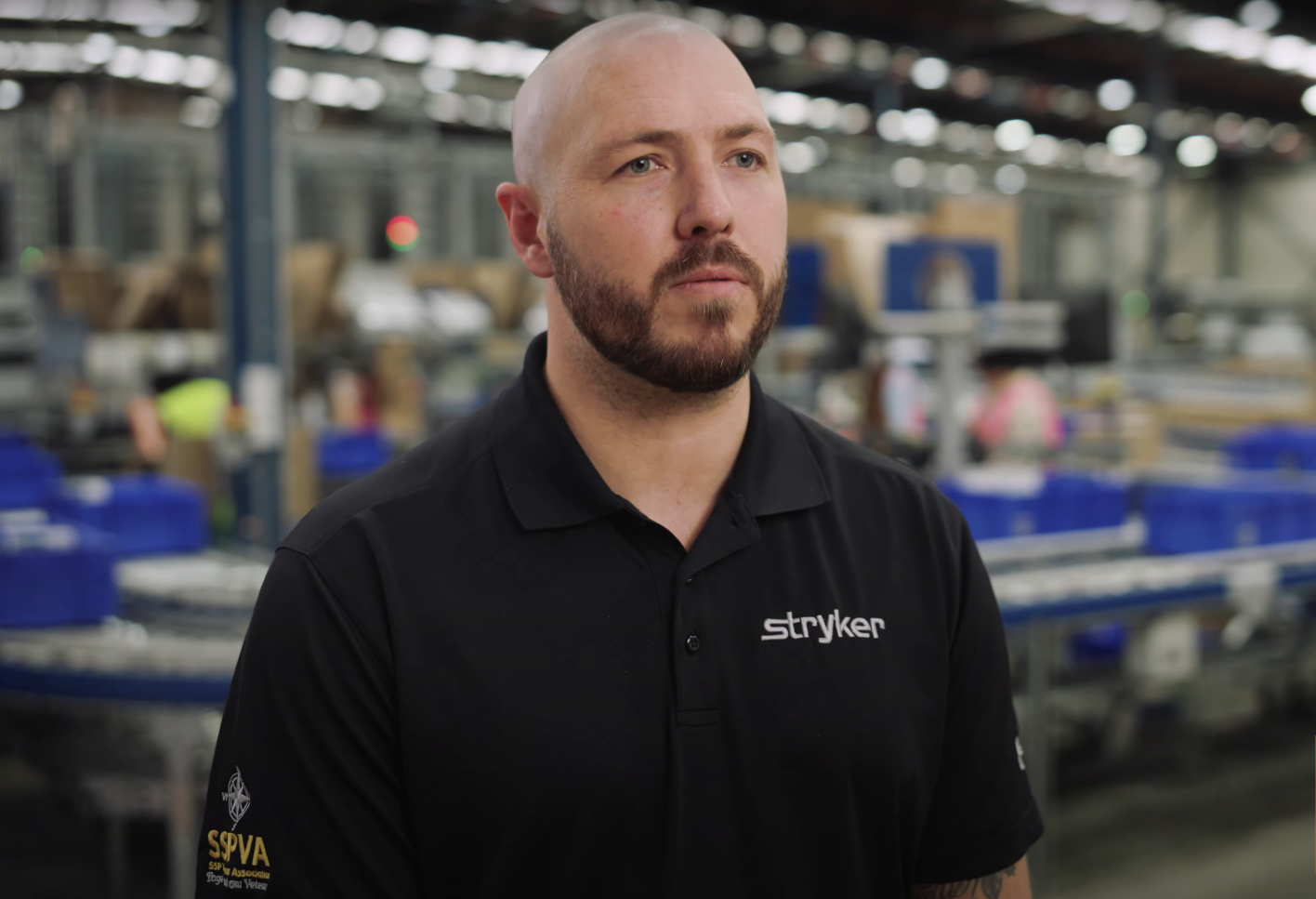 A bearded man in a black Stryker polo shirt stands in an factory setting