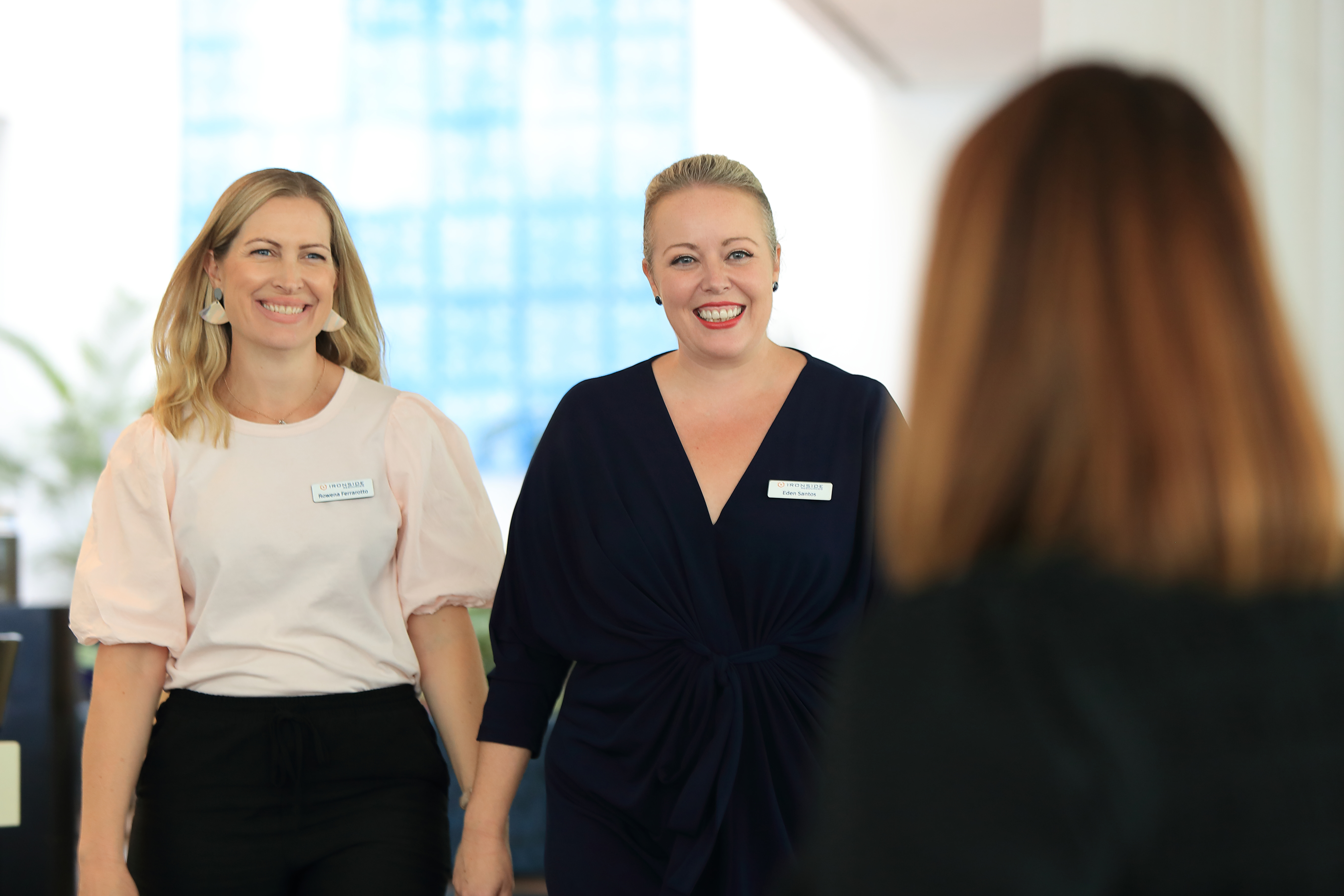 In the foreground, the back of a brunette person, in the background, two smiling women