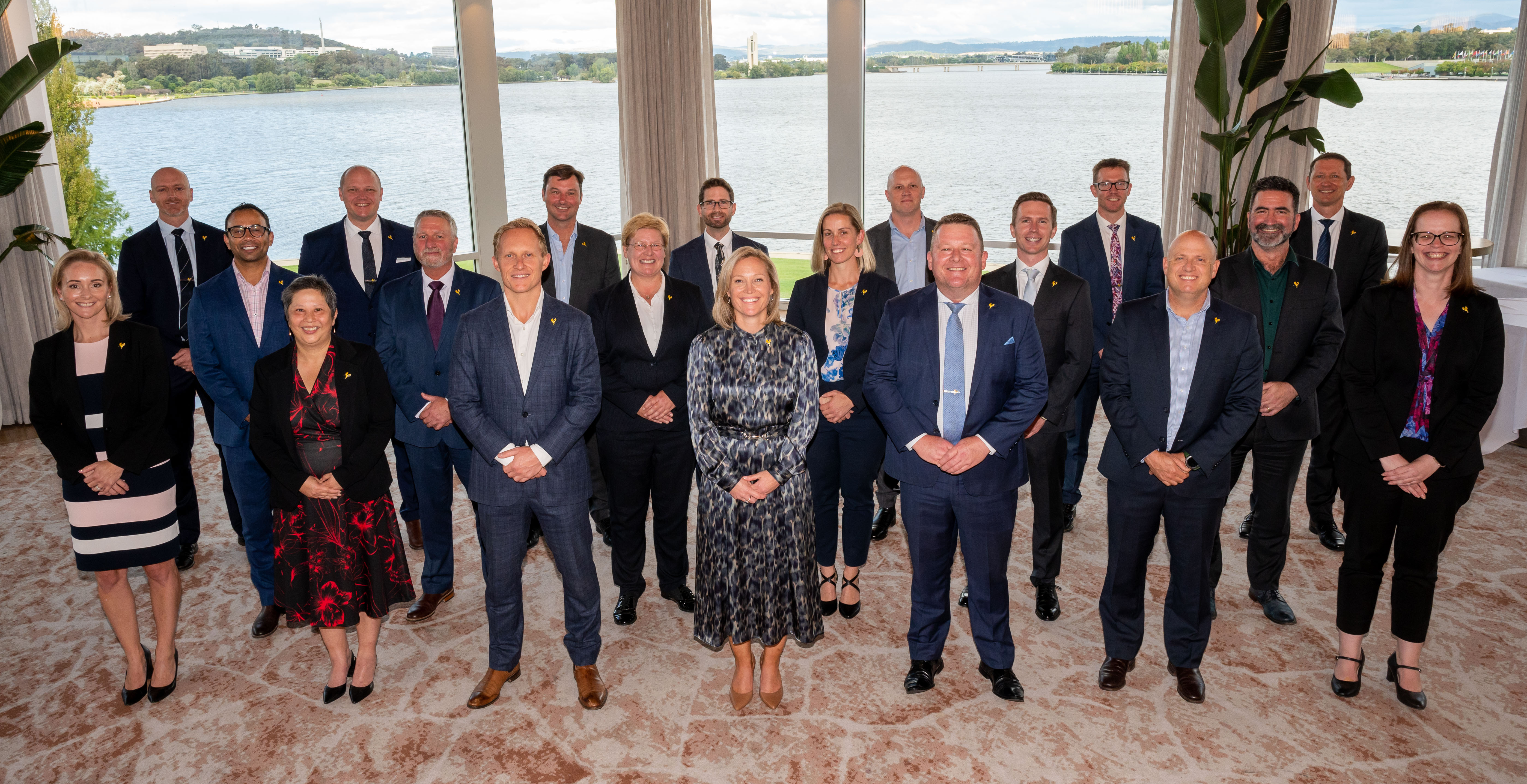 Three rows of people in business casual dress, standing in front of a window with a lake view