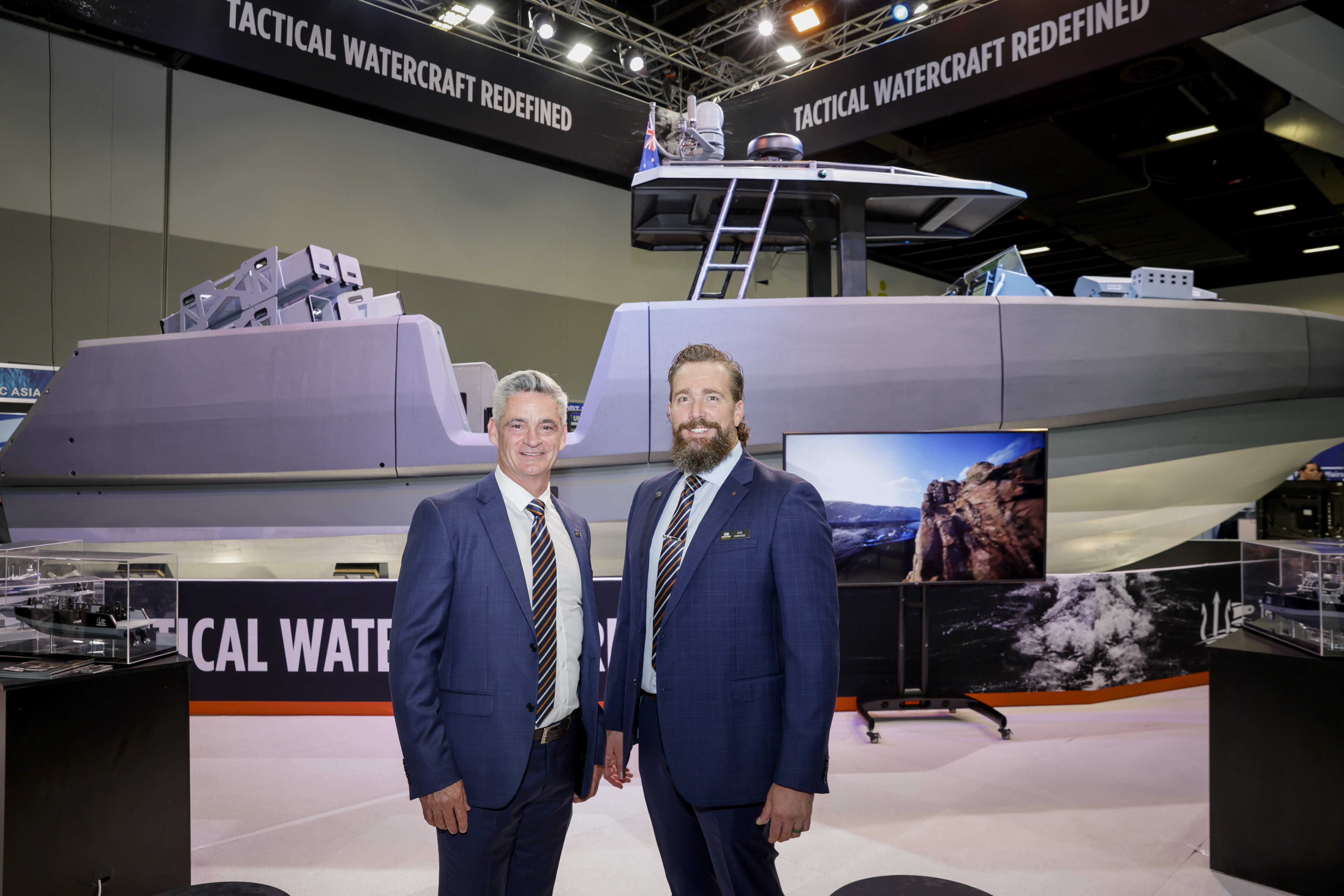 Two men in suits stand, smiling, in front of a full-size, display watercraft