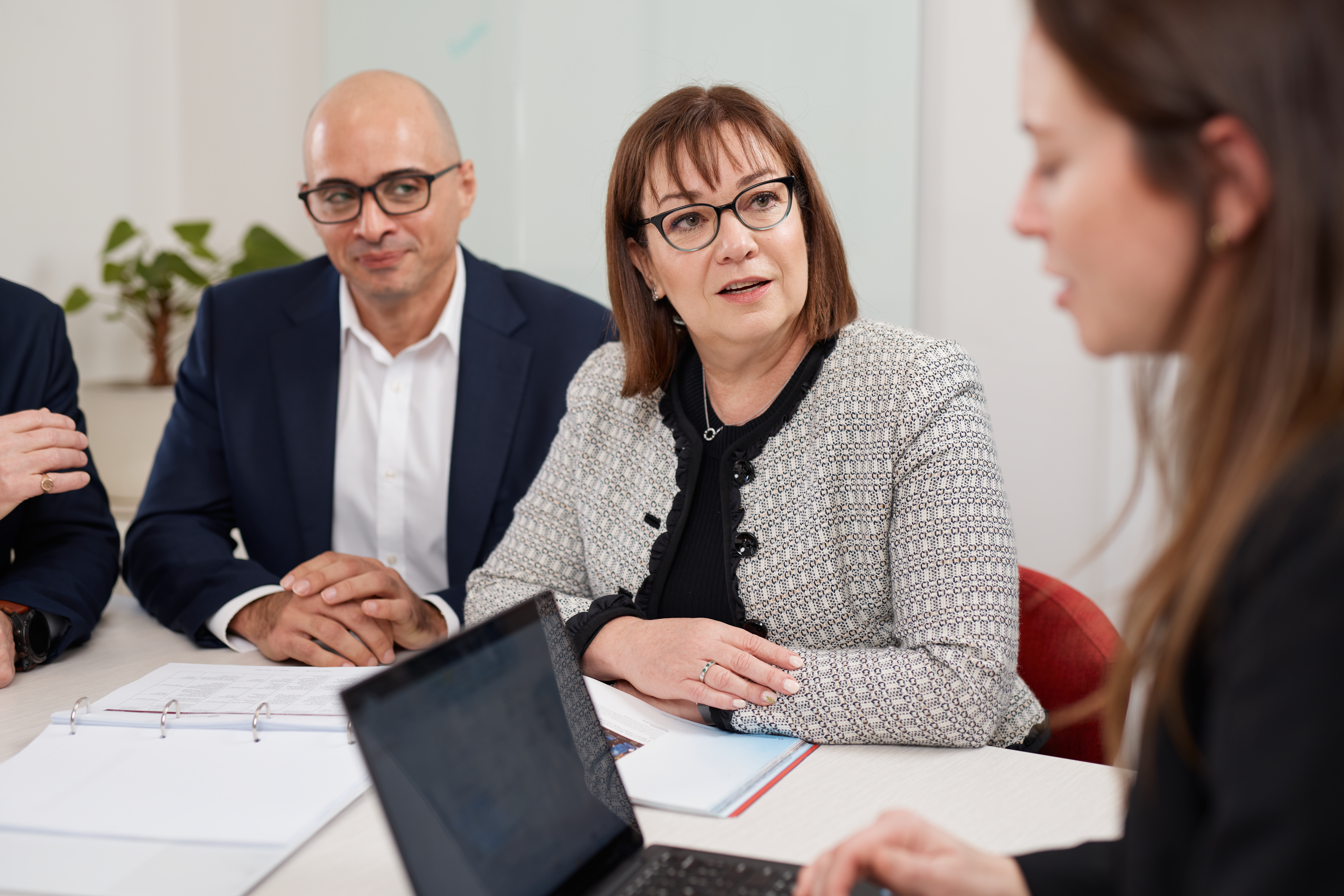 In focus, a woman and man in business attire look towards a woman, out of focus, in the foreground with a laptop.