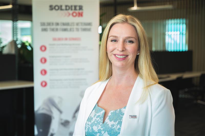 A blonde woman in a white blazer and blue and white top stands in front of a Soldier On banner