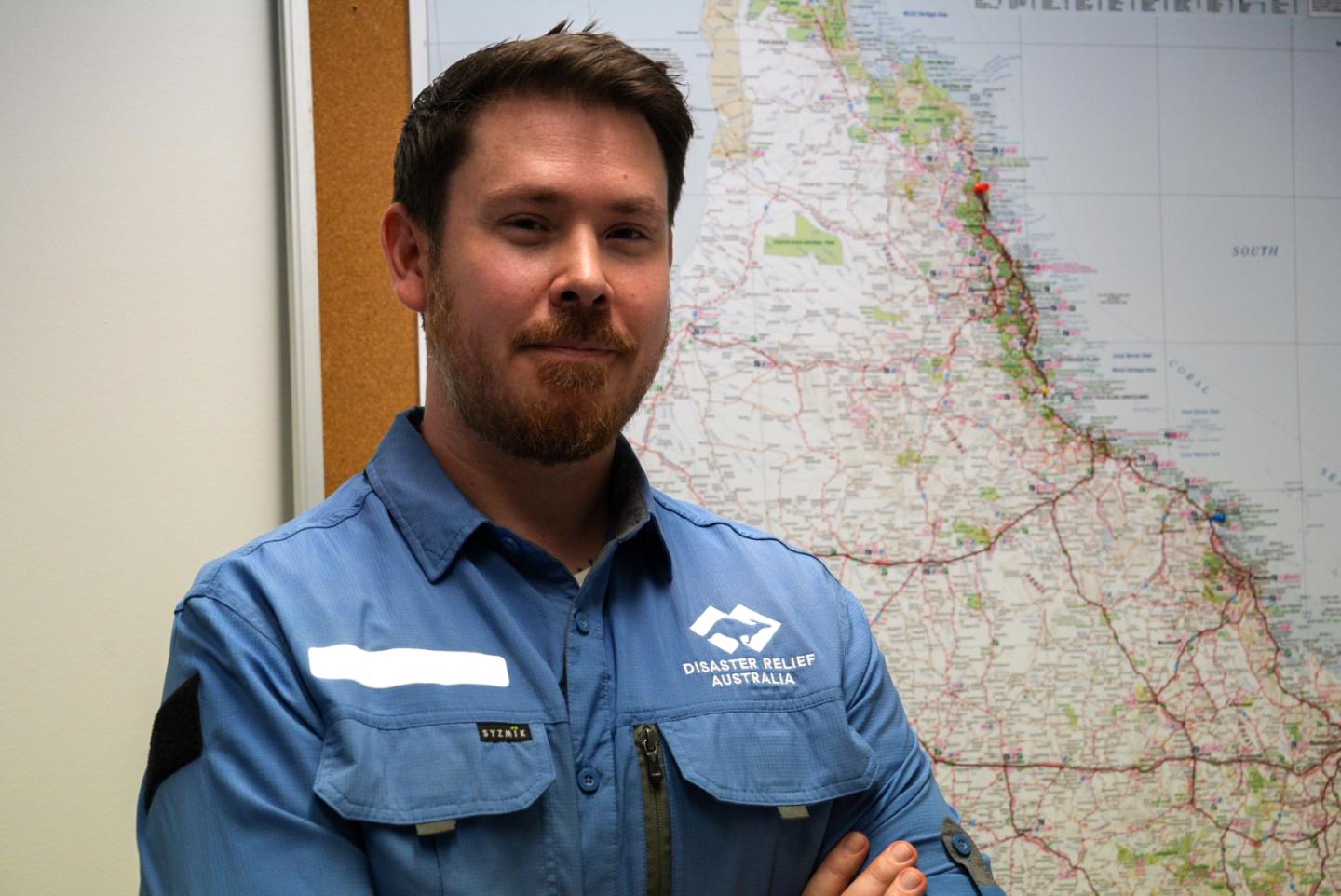 A dark-haired man in a blue DRA logo shirt stands in front of a map of Queensland