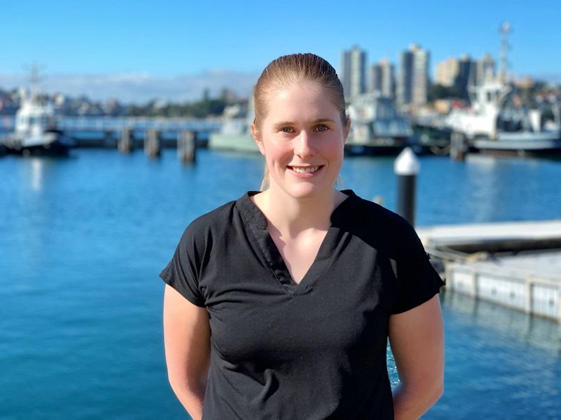 Headshot of a light-haired, smiling woman in a black top in front of a waterfront scene