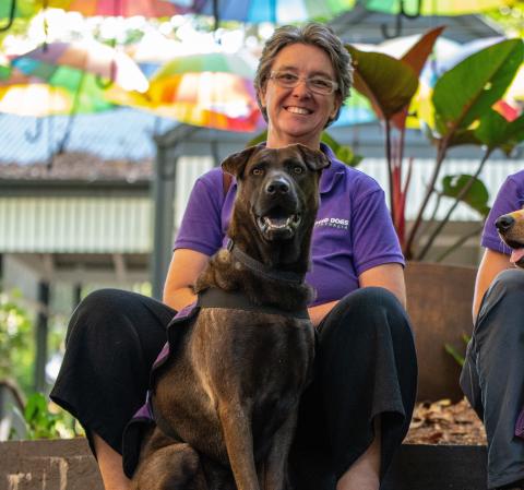 A woman in a purple polo shirt sits with a dark coloured dog