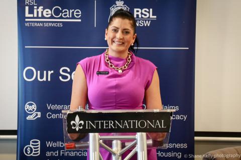 A dark-haired woman in a bright pink dress stands at a lectern