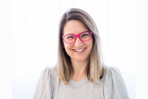 Headshot of a smiling blonde woman in pink glasses and a grey top